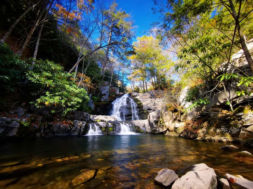 A stunning, wide-angle shot of Hawk Falls cascading into a crystal clear pool surrounded by lush rhododendrons and vibrant autumn foliage.